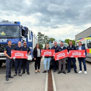 Eine Gruppe von neun Personen steht vor Einsatzfahrzeugen des Technischen Hilfswerks (THW) und anderer Rettungsdienste. Sie halten symbolische Spendenschecks in den Händen. Die Schecks zeigen unterschiedliche Beträge: 6.000 €, 5.250 €, 3.500 € und 2.500 €. Im Hintergrund sind ein großes blaues THW-Fahrzeug sowie ein Krankenwagen zu sehen. Die Gruppe lächelt, der Himmel ist bewölkt.