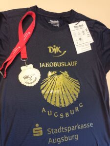 Ein T-Shirt mit einem Anhänger und einem Medaillenband. Das T-Shirt ist dunkelblau mit goldenen Aufdrucken: „DJK JAKOBUSLAUF AUGSBURG“ und einem Muschelsymbol. Das Medaillenband ist rot und trägt das Logo von Stadtsparkasse Augsburg. Die Medaille zeigt das Logo des Jakobuslaufs und die Zahl „25“, was auf den 25. Jubiläumslauf hinweist.