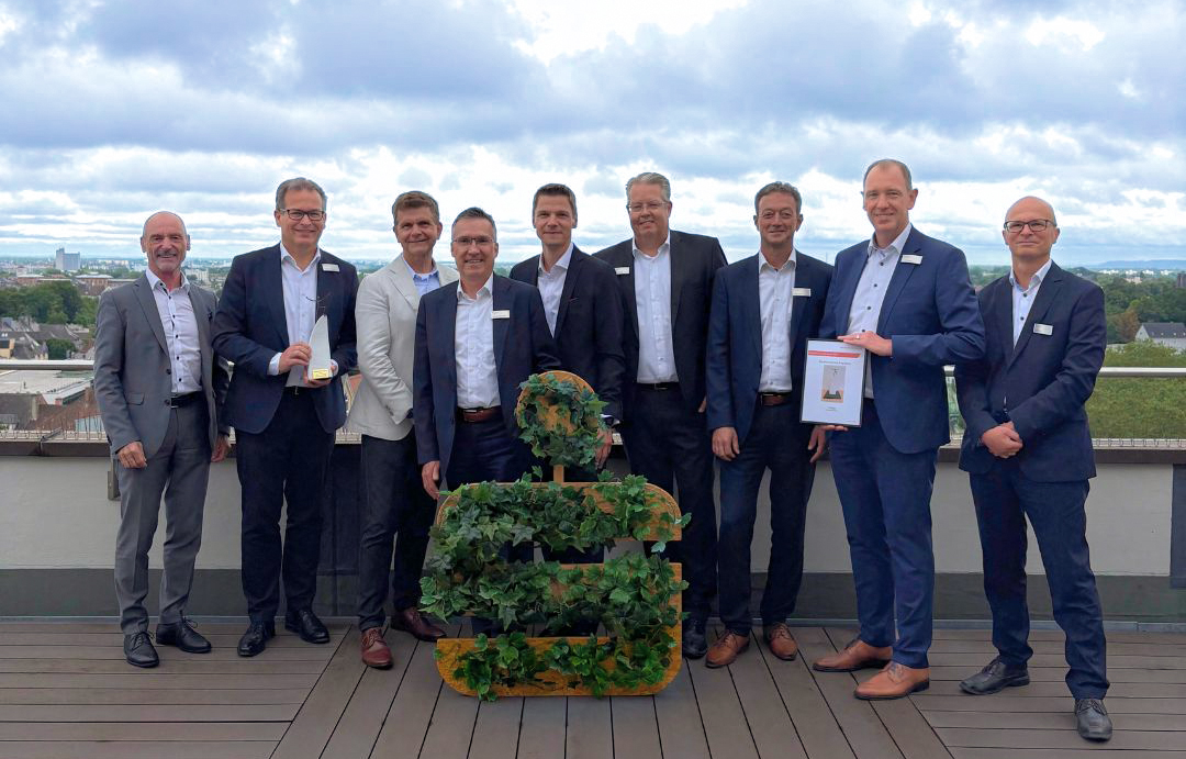 Neun Männer in Anzügen stehen auf einer Dachterrasse. Zwei von ihnen halten eine Auszeichnung und eine Urkunde in den Händen. Vor der Gruppe steht ein großes Sparkassen-Logo, das mit grünen Pflanzen dekoriert ist.