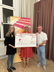Drei Personen stehen in einem Raum und halten gemeinsam einen symbolischen Spendenscheck der Stadtsparkasse Augsburg über 750 Euro für das Junge Theater Augsburg. Im Hintergrund ist ein Banner mit farbigen Streifen zu sehen.