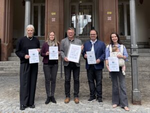 Fünf Personen stehen vor einem historischen Gebäude mit steinernen Treppen und halten Urkunden der Kinderstiftung Schwaben in den Händen. Eine Frau rechts hält zusätzlich ein kleines Kind auf dem Arm.