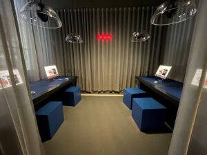 Eine moderne Medienstation in einem Raum mit minimalistischer Einrichtung. Zwei Computerarbeitsplätze sind mit blauen Würfeln als Stühlen und Tablets ausgestattet. Die Wände sind mit grauen Vorhängen bedeckt, und über den Arbeitsplätzen hängen große, moderne Lampen. Im Hintergrund ist ein rotes „DIGITAL“-Schild sichtbar.