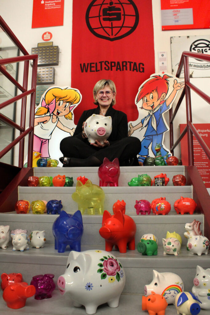 Cornelia Kollmer, stellvertretende Vorstandsvorsitzende der Stadtsparkasse Augsburg, blickt im historischen Archiv der SSKA gemeinsam mit Sparschweinen aus verschiedenen Dekaden auf den Weltspartag und die Kindersparwoche vom 30.10. bis 07.11.2025.