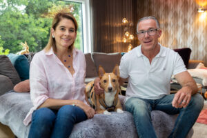 Ein Paar sitzt auf einem Sofa in einem gemütlichen Raum, mit einem Hund auf dem Schoß der Frau. Beide lächeln freundlich in die Kamera. Der Raum hat moderne Möbel und große Fenster, durch die Grünpflanzen und Sonnenlicht zu sehen sind.