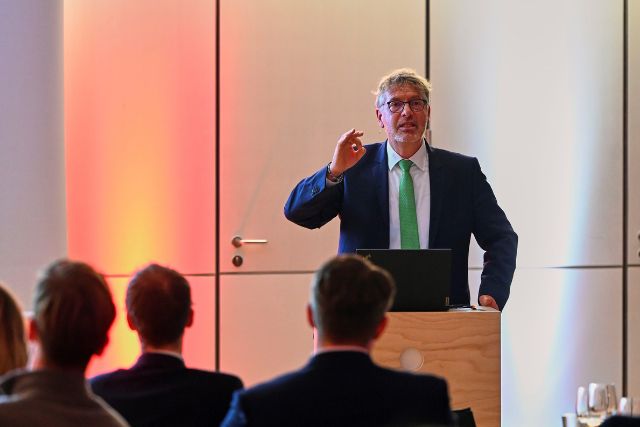 Das Bild zeigt Philipp Vorndran, der vor einem Publikum eine Präsentation hält. Er trägt einen Anzug mit grüner Krawatte und gestikuliert mit der Hand. Im Hintergrund sind sanfte Lichtakzente zu sehen.