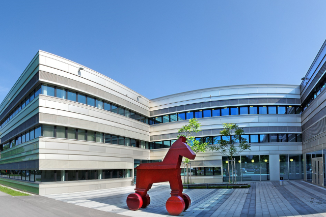 Moderne Architektur eines Bürogebäudes mit einer roten Skulptur eines Pferdes im Vordergrund, bei sonnigem Wetter.
