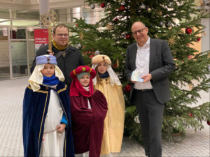 Die Sternsinger 2026, die in traditionellen Gewändern als Heilige Drei Könige vor einem Christbaum in der Schalterhalle der Stadtsparkasse Augsburg stehen. Herr Mahler von der Stadtsparkasse Augsburg übergibt ihnen Geldscheine.