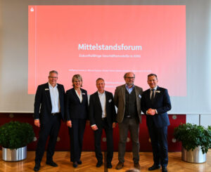 Vier Männer und eine Frau stehe von einer Präsentation der Stadtsparkasse Augsburg mit dem Text: Mittelstandsforum, zukunftsfähige Geschäftsmodelle in KMU.