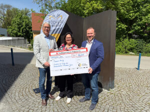 Drei Personen stehen draußen vor der Bücherei Mering und halten gemeinsam einen großen symbolischen Scheck über 706 Euro. Die Person in der Mitte trägt ein bunt gemustertes Oberteil, die beiden Personen links und rechts einen hellen bzw. dunkelblauen Blazer. Im Hintergrund ist ein Banner mit dem Logo der Bücherei Mering und Bäume zu sehen.