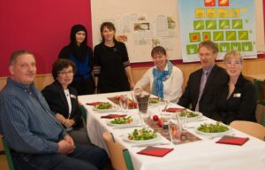 Sieben Erwachsene sitzen und stehen gemeinsam an einem festlich gedeckten Tisch mit Salat und Getränken. Die Aufnahme entstand in einem Gemeinschaftsraum mit Informationsplakaten an der Wand und zeigt eine gemeinsame Mahlzeit in freundlicher Atmosphäre.