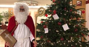 Ein als Nikolaus verkleideter Mann mit rotem Umhang, Bischofsstab und Geschenk steht neben einem festlich geschmückten Weihnachtsbaum. Am Baum hängen Wunschkarten, darunter liegen Geschenke in einer Filiale der Stadtsparkasse Augsburg.