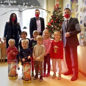 Sieben Kinder stehen lächelnd vor einem geschmückten Weihnachtsbaum und präsentieren Geschenke. Hinter ihnen stehen zwei Mitarbeitende der Stadtsparkasse Augsburg mit roten Masken und begleiten die Geschenkübergabe.