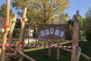 Ein neu gestalteter Kletter- und Spielbereich aus Holz ist mit bunten Luftballons geschmückt. In der Mitte hängt ein großes Plakat mit der Aufschrift ‚DANKE‘, umgeben von Kinderzeichnungen und Handabdrücken.