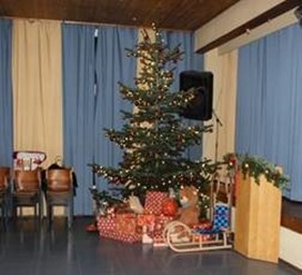 Ein geschmückter Weihnachtsbaum steht in einem festlich dekorierten Saal. Unter dem Baum liegen zahlreiche verpackte Geschenke, daneben stehen ein Holzschlitten und weitere weihnachtliche Dekoration.