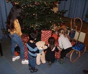 ier Kinder stehen vor einem geschmückten Weihnachtsbaum und betrachten die verpackten Geschenke darunter. Ein Kind hält einen Teddybären, daneben steht ein Holzschlitten