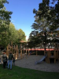 Ein weitläufiger Spielplatz mit Klettergerüsten, Rutsche und Sandfläche liegt unter großen Bäumen. Im Vordergrund stehen zwei Erwachsene mit einem Kind und blicken auf den Spielbereich.