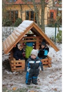 Mehrere Kinder in Winterkleidung spielen in einem kleinen Holzspielhaus auf einem leicht verschneiten Spielplatz. Ein Kleinkind steht davor, im Hintergrund sind Bäume und ein Gebäude zu sehen.