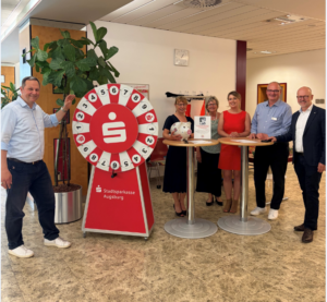 Sechs Personen stehen in einer Filiale der Stadtsparkasse Augsburg neben einem roten Glücksrad. Einige präsentieren eine Sparschwein-Spendenbox und eine Urkunde. Die Gruppe lächelt in die Kamera und steht an Stehtischen im Beratungsbereich.