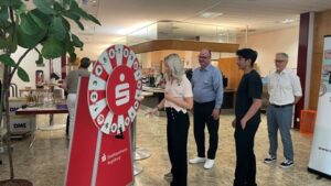 Eine Frau dreht an einem roten Glücksrad der Stadtsparkasse Augsburg, während drei weitere Personen gespannt daneben stehen und zusehen. Die Szene spielt im Beratungsbereich einer Filiale der Stadtsparkasse Augsburg.