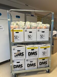 Ein Rollwagen ist mit mehreren Kartons und zahlreichen gepackten Lebensmitteltüten beladen. Die Tüten stehen auf den Kartons und sind für eine Verteilaktion vorbereitet. Im Hintergrund befinden sich Kühlschränke und ein Lagerraum.