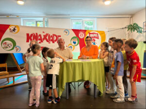 Kinder versammeln sich um Sandra Peetz-Rauch und Norbert Schad vor dem KNAXIADE-Banner und schauen sie aufmerksam an.