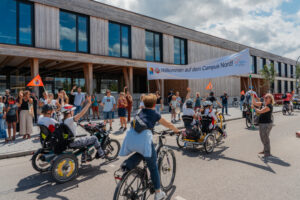 Eine Gruppe von Radfahrenden, darunter Menschen mit und ohne Behinderung, fährt gemeinsam an einem modernen Holzgebäude vorbei. Zahlreiche Zuschauerinnen und Zuschauer begrüßen den Fahrradkonvoi unter einem Banner mit der Aufschrift ‚Willkommen auf dem Campus Nord‘ und winken den Teilnehmenden zu.