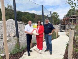 Drei Personen stehen im Zoo Augsburg vor einer Baustelle und präsentieren symbolisch eine Spende über 2.500 Euro. Im Hintergrund sind ein neu gestaltetes Gehege, Bäume und ein Gebäude im Bau zu sehen.