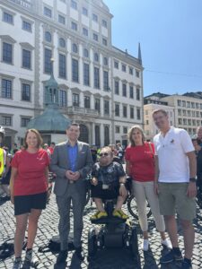 Fünf Personen stehen auf dem Augsburger Rathausplatz vor dem Rathaus und lächeln in die Kamera. In der Mitte sitzt ein Mann in einem Elektrorollstuhl. Im Hintergrund sind zahlreiche Teilnehmende einer Veranstaltung sowie Fahrräder zu sehen.