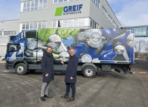Zwei Männer stehen vor einem bunt gestalteten Wasserstoff-Lkw von GREIF Mietwäsche und lächeln in die Kamera. Im Hintergrund ist das Firmengebäude mit dem GREIF-Logo zu sehen.